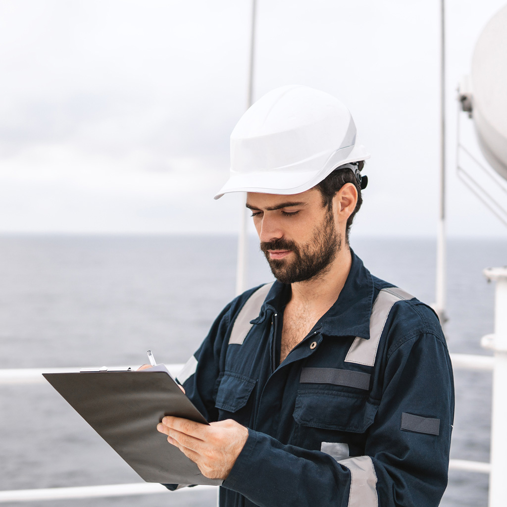 Marine engineer in safety gear reviewing documents onboard, representing marine insurance plans for ships & cargo.